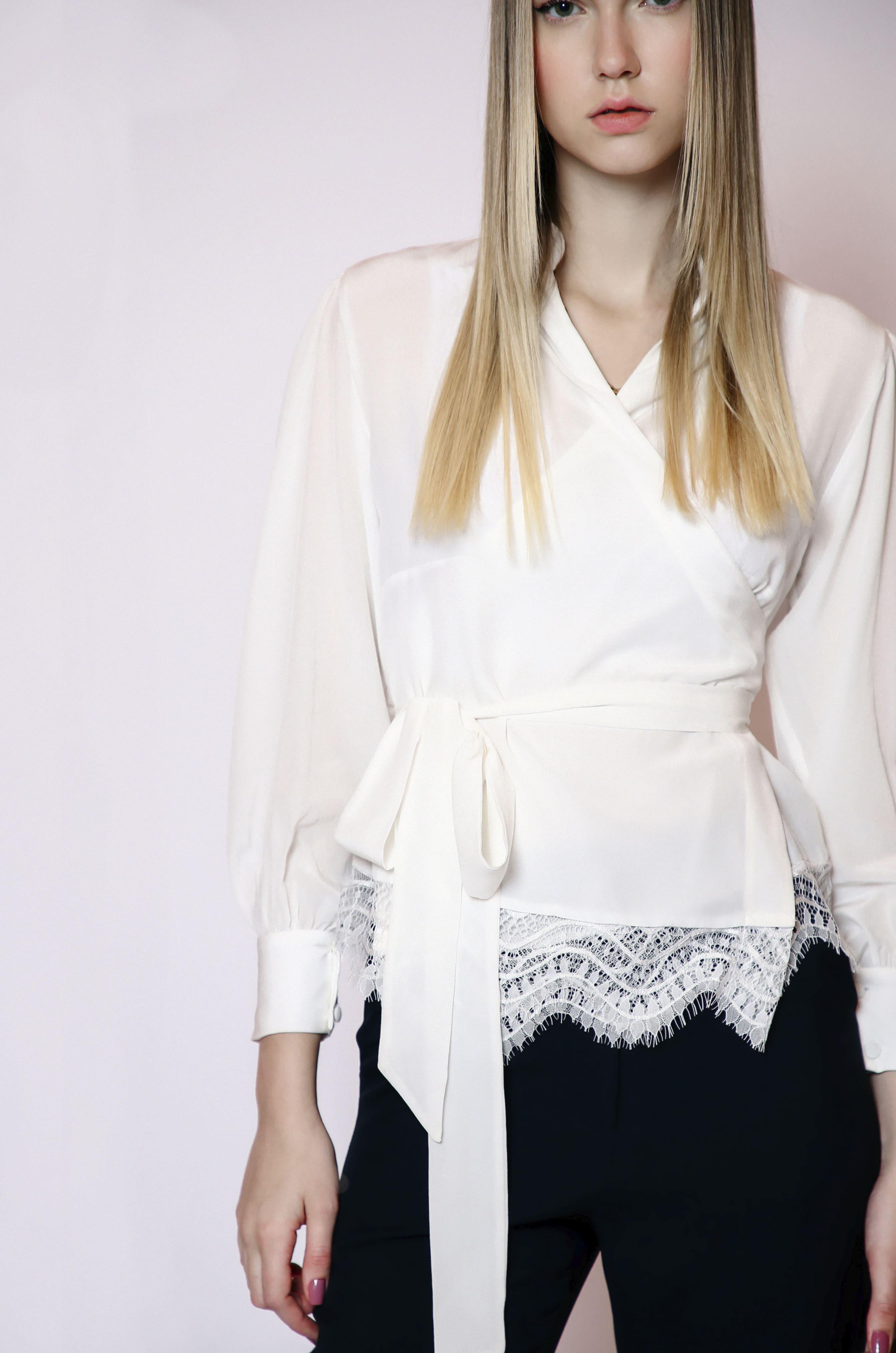 A woman poses against a plain backdrop wearing LETHICIA BRONSTEINs Cache-Coeur Blouse Auzira in off-white silk with lace details and a tie belt, paired with black pants. Her long hair is straight and loose.