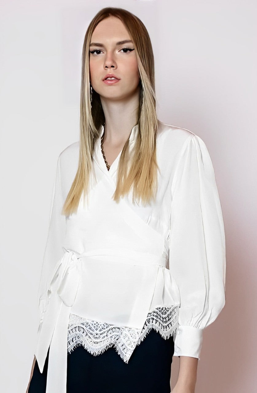 A woman with long straight hair wears the LETHICIA BRONSTEIN Cache-Coeur Blouse Auzira in Off-White Silk, featuring lace trim and a waist tie. She stands against a plain background, looking forward.