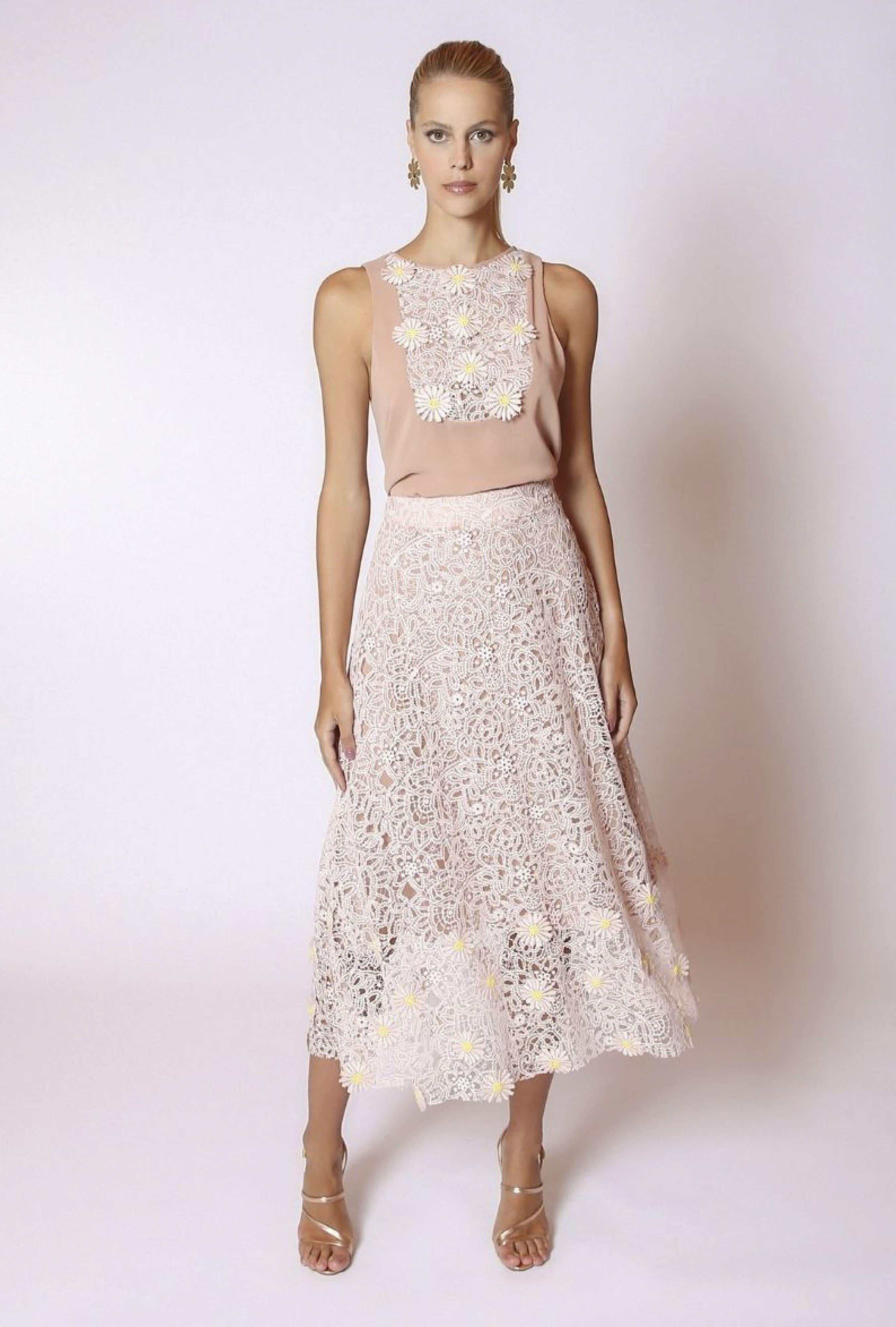 A woman in a LETHICIA BRONSTEIN Barbara sleeveless blouse in daisy rose georgette with guipure and a matching lace floral skirt stands against a plain backdrop. Her clear high-heeled sandals enhance her look, with styled back hair accessorized with earrings.