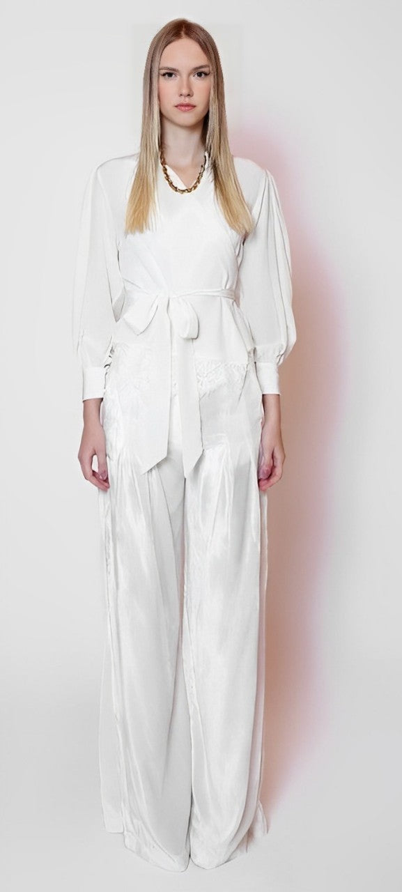 A person with long hair stands against a plain background wearing a LETHICIA BRONSTEIN Cache-Coeur Blouse Auzira in off-white silk. The blouse, featuring a belted waist and wide-leg pants, is complemented by their natural makeup, neutral expression, and an elegant necklace.