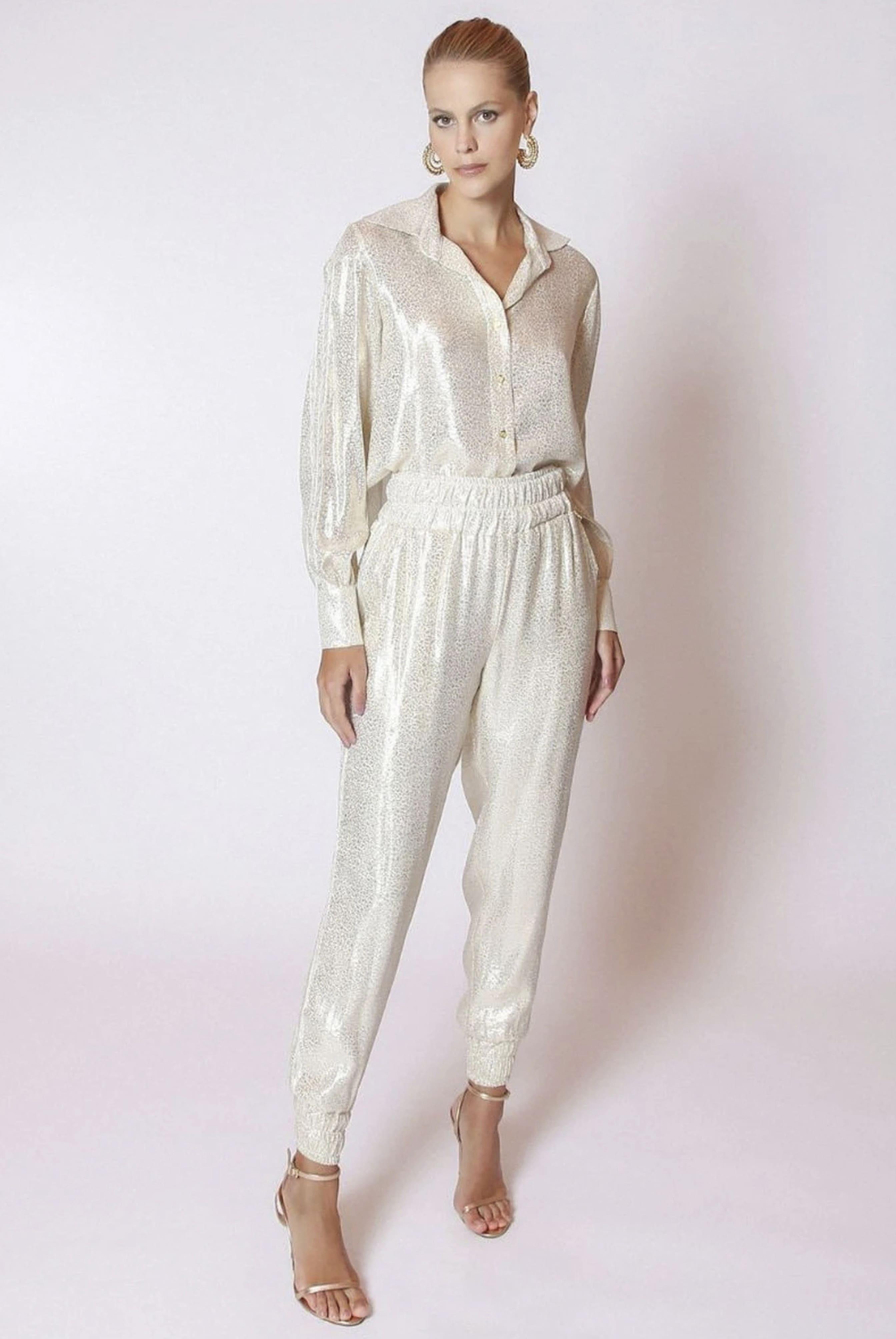 A woman confidently poses in LETHICIA BRONSTEINs Bibiana Foil Pajama Pants with Lace in Off-White, complemented by hoop earrings and high-heeled sandals, against a plain white background.