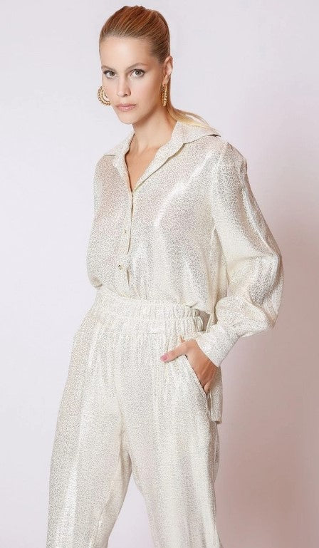 A person with long hair poses against an off-white background, wearing a shimmering metallic long-sleeve blouse featuring lace accents, along with LETHICIA BRONSTEIN Bibiana Foil Pajama Pants. One hand is casually placed in their pocket.