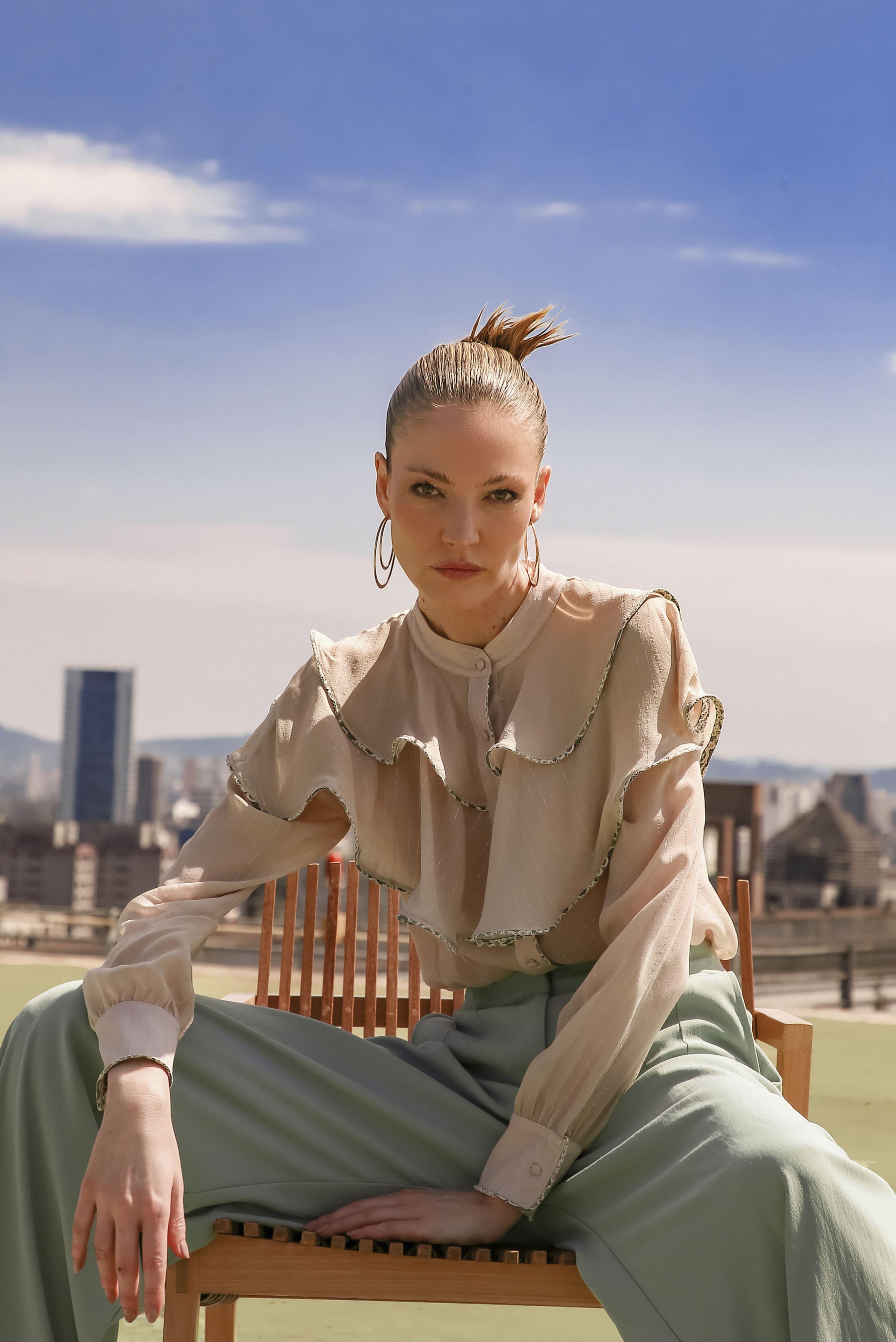 Sitting confidently on a rooftop bench, a person wears the LETHICIA BRONSTEIN Ana Ruffled Shirt in Natural Organza and light green pants, their hair in a top knot against the clear blue sky and cityscape backdrop.