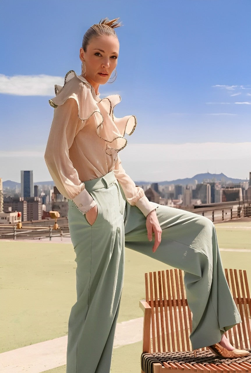 A woman stands confidently on a rooftop, cityscape behind her. Shes wearing a LETHICIA BRONSTEIN Ana Ruffled Shirt in Natural Organza with wide-leg, high-waisted mint green pants. Her hair is pulled back as she poses next to a wooden bench beneath a clear blue sky.