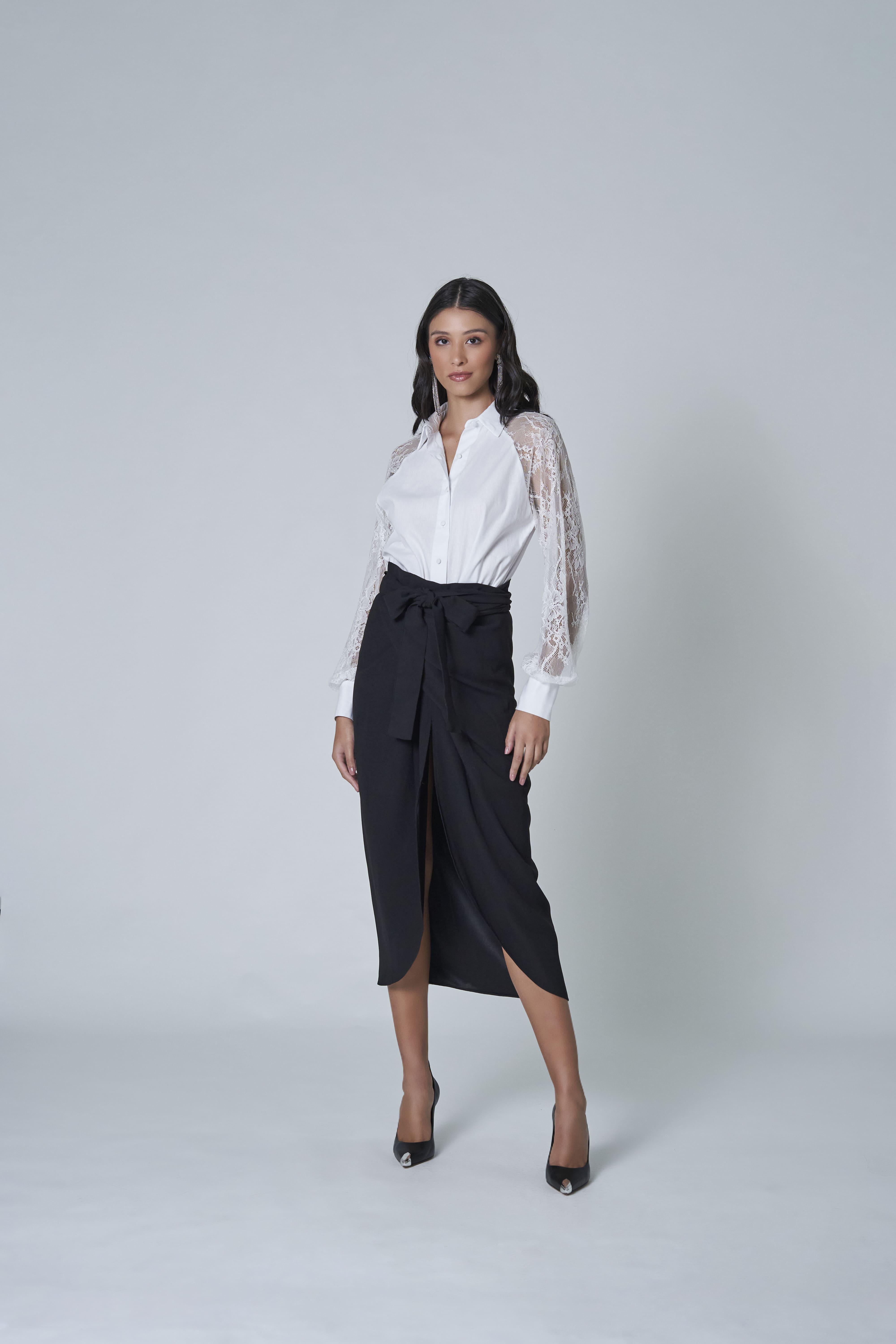 In a studio against a gray background, a woman confidently wears the stunning LETHICIA BRONSTEIN Amy Tricoline and Lace Shirt in white, paired with a high-waisted black skirt tied at the waist and black heels.