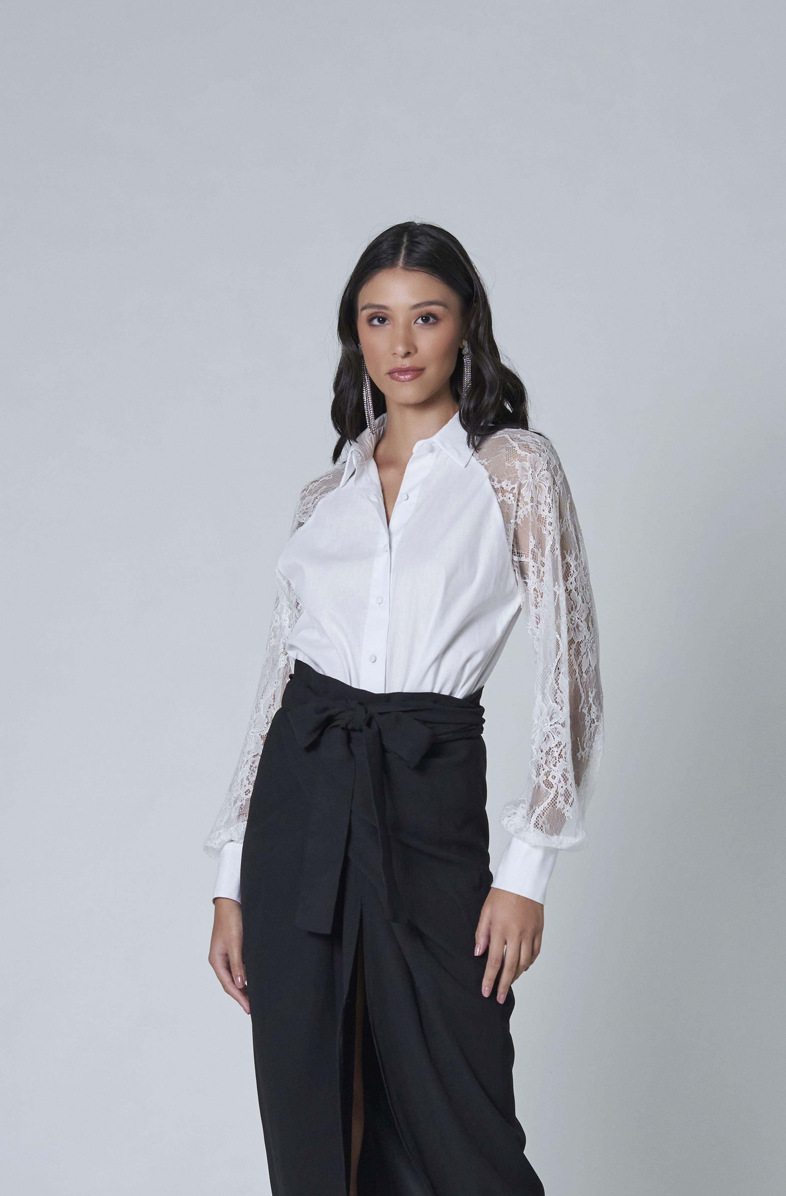 Against a plain backdrop, a woman models the LETHICIA BRONSTEIN Amy Tricoline and Lace Shirt in White. Her long dark hair cascades as she gazes at the camera, paired with a black skirt featuring a bow for an elegant look.