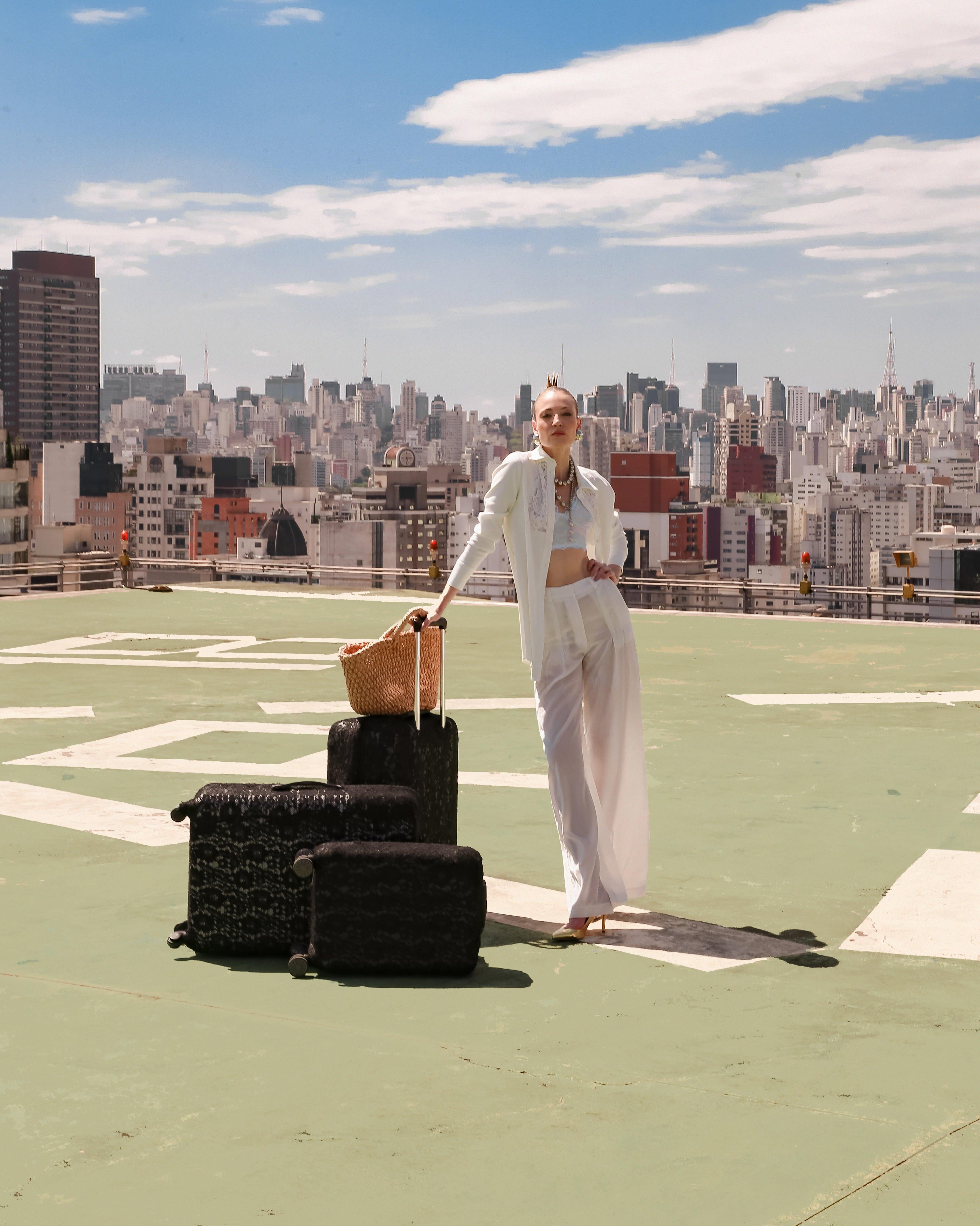 A person stands confidently on a rooftop helipad with a cityscape backdrop. Theyre wearing the LETHICIA BRONSTEIN Long Crepe and Lace Shirt Aida in Off-White, along with sunglasses, near a large straw bag atop three black suitcases. The sky is partly cloudy.